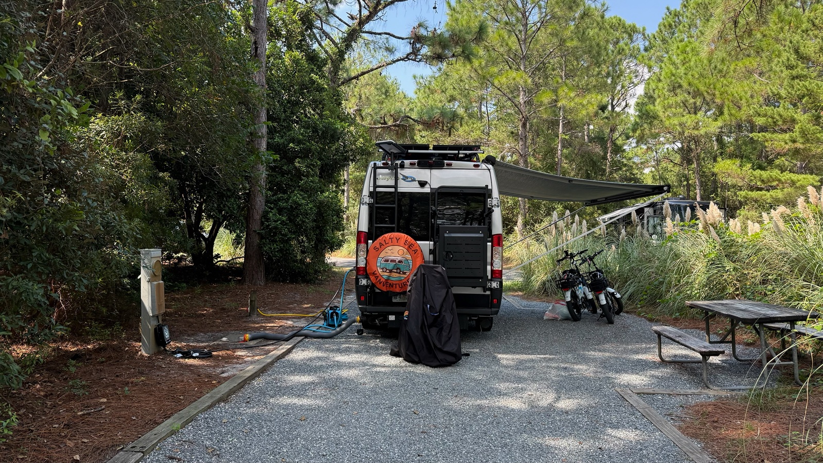 Rear of a camper van with awning extended and bikes at a wooded campsite