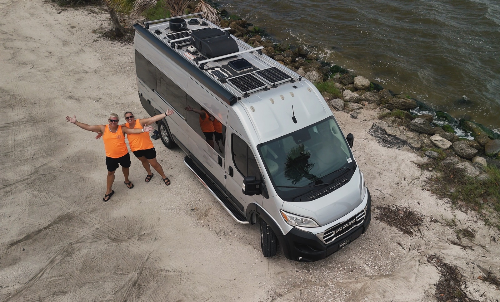 Two travelers waving beside a silver camper van by the water